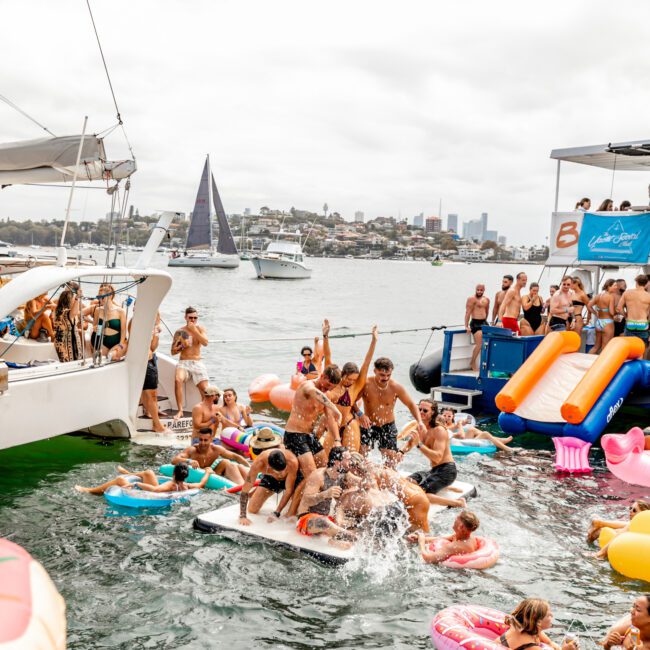 A lively group of people partying on and around a boat in Sydney Harbour. Many are on floaties in the water, while some dance on a floating platform. Colorful inflatables add to the fun, with another boat and several sailboats in the background under a cloudy sky, reminiscent of The Yacht Social Club's famed Boat Parties Sydney.