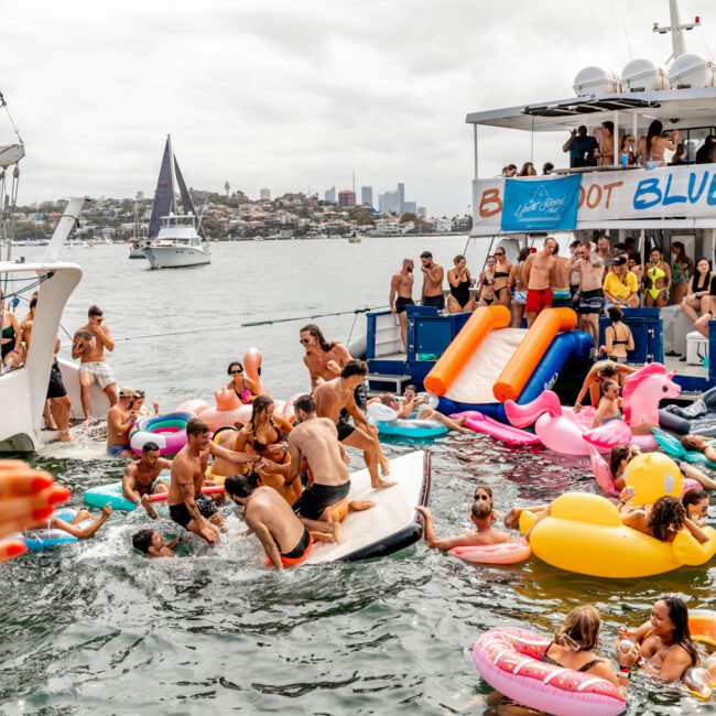 A lively boat party with people swimming among colorful inflatable floats. The backdrop features yachts and a coastal city skyline under a cloudy sky. A large boat with a "Boot Blue" banner is docked, and participants relish music and dancing, enjoying The Yacht Social Club Event Boat Charters.