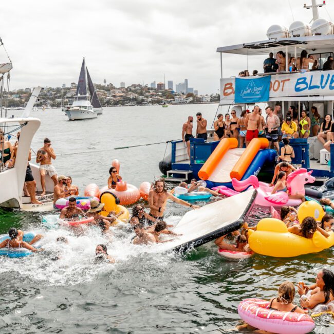 A lively boat party hosted by The Yacht Social Club Sydney Boat Hire features people enjoying the water on colorful inflatables. Two boats are docked side by side, with many gathered on one boat and in the water, using slides to connect to the sea. The background showcases a stunning coastal cityscape.