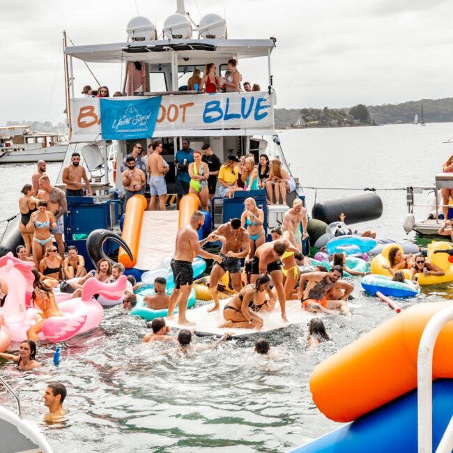 A lively gathering on a large boat named "Big Boy Blue" features numerous people swimming and using inflatable toys in the water. The festive scene, hosted by The Yacht Social Club Sydney Boat Hire, includes attendees on a slide surrounded by colorful pool floats. In the background, a calm lake and distant land complete the picturesque setting.