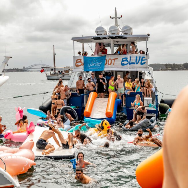 A lively scene of people enjoying a party on a yacht and in the surrounding water. The yacht has a "Yacht Social Club" and "Boot Blue" sign. Numerous inflatable toys, including animals and tubes, are in the water. The event is part of The Yacht Social Club Sydney Boat Hire offerings. The sky is overcast.