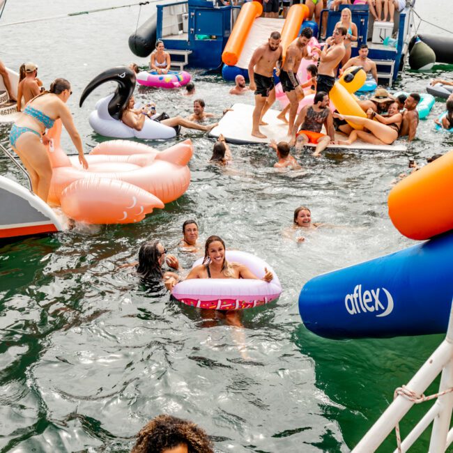 A lively gathering of people enjoying a day on the water with colorful inflatables, floating near anchored boats. Some individuals are in the water while others are on the boats or inflatables, engaging in conversations and having a good time under mostly overcast skies. Experience this at The Yacht Social Club Sydney Boat Hire.