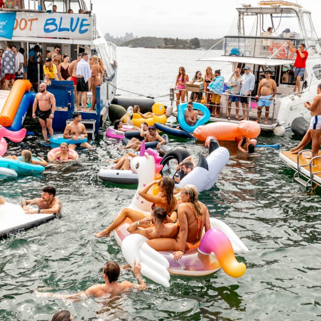 A lively scene of a boat party with numerous people enjoying themselves on inflatable floats in the water. Several boats, part of the Boat Parties Sydney The Yacht Social Club, are anchored close together, with partygoers socializing onboard and in the water. Some use vibrant floats shaped like unicorns and donuts.