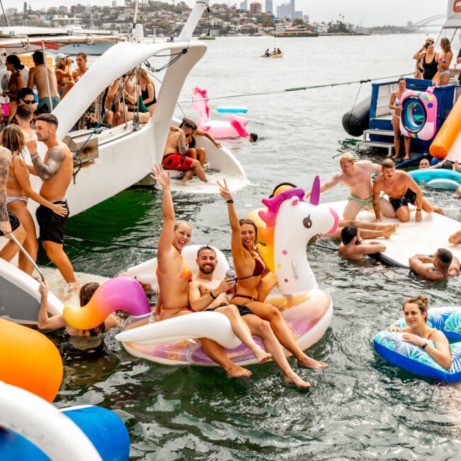 A lively group of people are enjoying a boat party on the water. Three on a large inflatable unicorn are smiling and raising their arms. Others swim, float on inflatables, and socialize on boats in the background at The Yacht Social Club Event Boat Charters. The sky looks overcast.