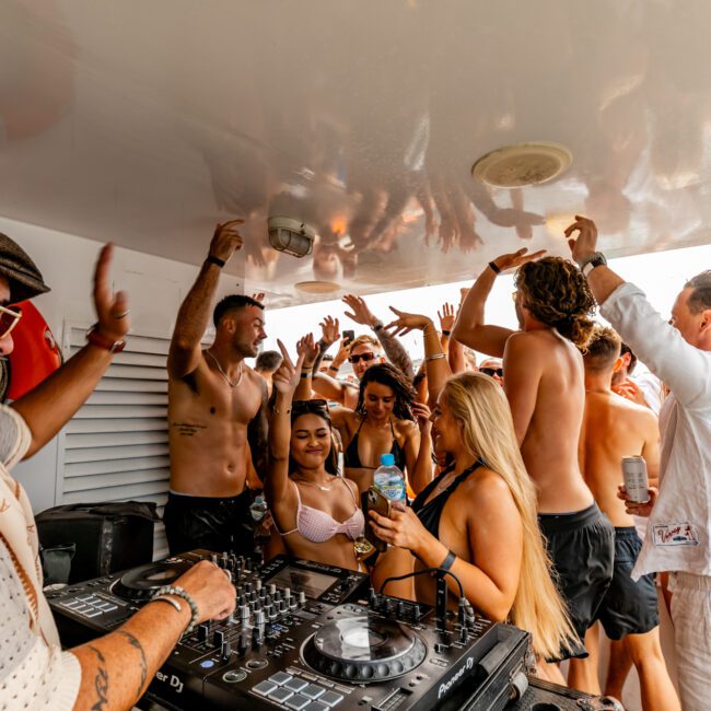 A DJ wearing headphones and sunglasses performs on a boat, surrounded by a lively crowd of young adults dancing and enjoying the music. The group includes men and women in swimwear, with some holding drinks, against the backdrop of a sunny, blue sea at The Yacht Social Club Event Boat Charters.