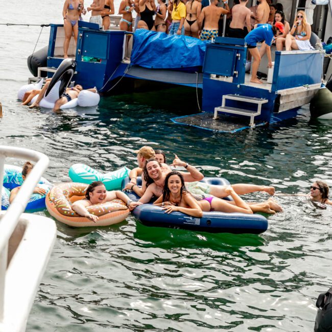 A group of people is having fun on floats in the water near a boat named "Barefoot Blue" by Sydney Harbour Boat Hire The Yacht Social Club. Additional people are on the boat, socializing and enjoying the sunny day, suggesting a lively, festive atmosphere on the waterfront.