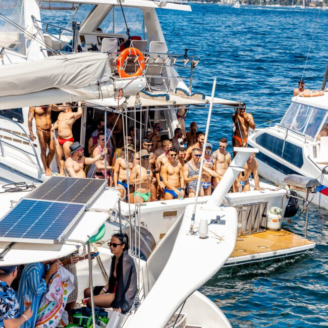 A lively group of people enjoying a sunny day on yachts during The Yacht Social Club Event Boat Charters. The boats are anchored close together on the water, with people wearing swimsuits and sunglasses, socializing, dancing, and taking photos. The city skyline and other boats are visible in the background.