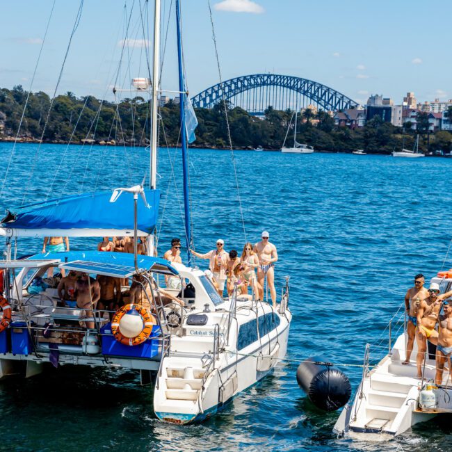 A group of people enjoying a sunny day on yachts in a blue body of water, with a large steel arch bridge visible in the background. Some are relaxing on the boats, while others are standing and chatting. Trees and city buildings can be seen in the distance, highlighting the fun experience offered by Sydney Harbour Boat Hire The Yacht Social Club.