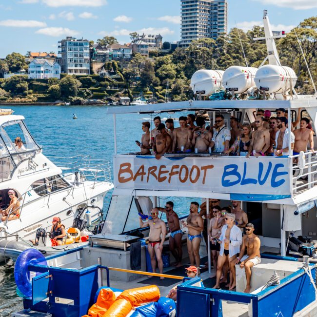 A group of people dressed in swimwear are gathered on a boat named "Barefoot Blue" on the water, enjoying a Yacht Social Club event. Another smaller boat is nearby. The backdrop features a scenic waterfront with modern buildings nestled among trees. Inflatable pool toys are seen on the boat's deck.