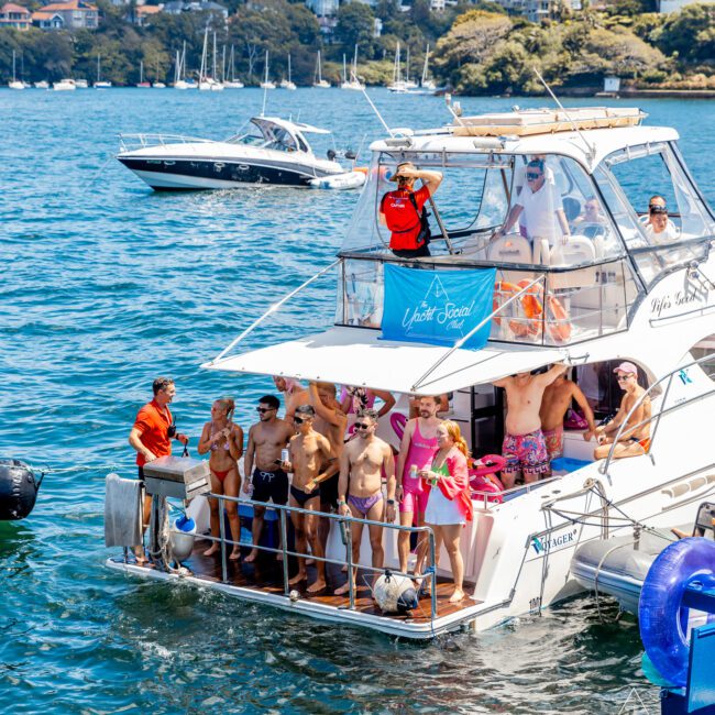 A group of people standing on the back deck of a boat in a picturesque harbor, wearing swimwear and smiling. The boat is anchored near other boats, with a cityscape and greenery in the background. Some are waving, with a logo reading "The Yacht Social Club Sydney Boat Hire.