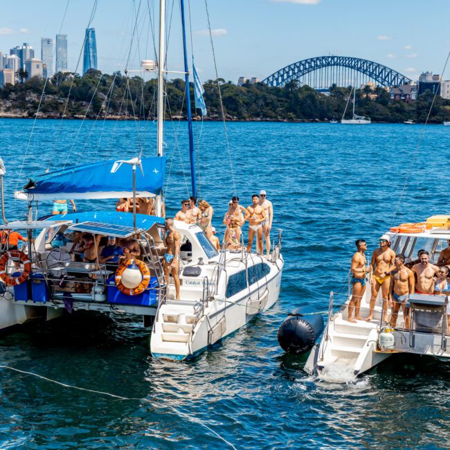 A lively group of people in swimsuits are partying on two catamarans on a body of water. The backdrop features a city skyline with high-rise buildings and a large arched bridge. It's a sunny day with clear blue water, and the mood is festive, courtesy of The Yacht Social Club Sydney Boat Hire.