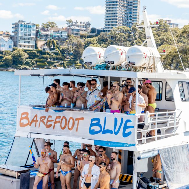 A large group of people in swimwear are gathered on a two-level boat with banners reading "Barefoot Blue." The Yacht Social Club Event Boat Charters is cruising on a sunny day, surrounded by lush green trees and shoreline buildings in the background. Ideal for Boat Rental and Parties Sydney The Yacht Social Club.