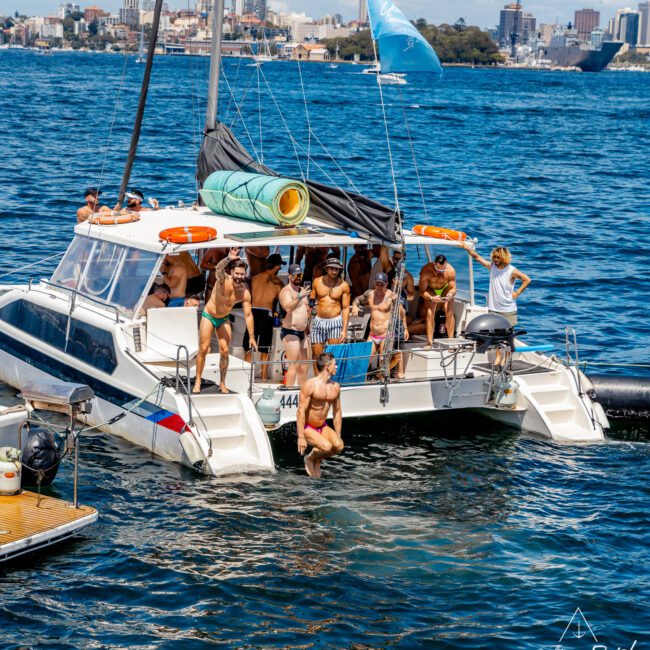 A group of people in swimwear are gathered on the deck of a catamaran. Some are sitting while others stand near the back. One person is jumping off into the water, and the boat sails on a large body of water with a cityscape visible in the background, showcasing The Yacht Social Club Sydney Boat Hire.