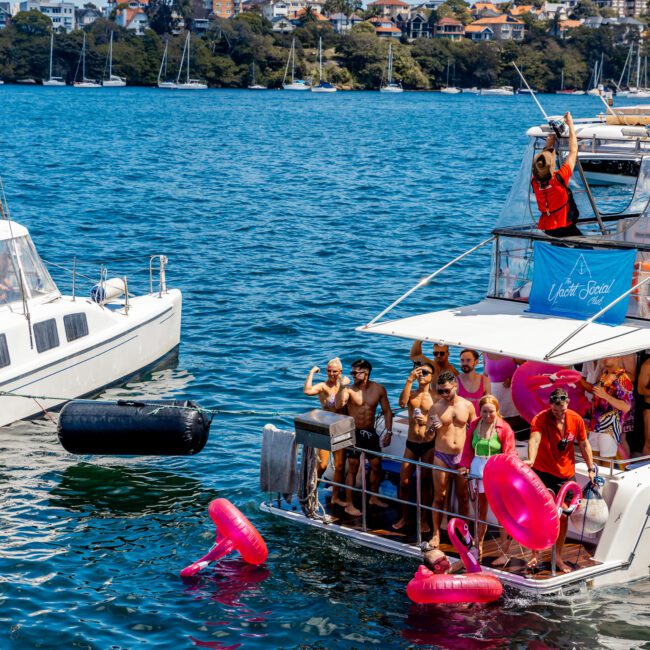 A group of people partying on the deck of a yacht, surrounded by water and a coastal cityscape in the background. Some are holding drinks and inflatable flamingos float nearby. Another vessel from The Yacht Social Club Sydney Boat Hire is anchored close by. The weather is sunny and clear.