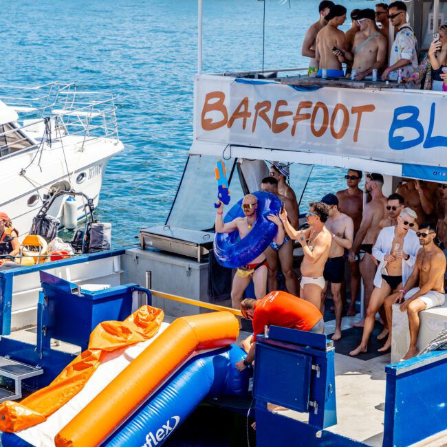 A group of people in swimwear enjoys a sunny day on a docked boat with a "Barefoot" banner. One person with an inflatable ring and water guns stands near an inflatable slide that descends into the water. Others are gathered, chatting, and having fun on the boat at The Yacht Social Club Event Boat Charters.