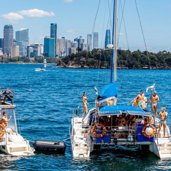 A group of people enjoy a sunny day on two sailboats anchored close together in a bustling harbor, with a modern city skyline visible in the background. Some individuals are sunbathing, while others are in the water or preparing to swim. This scene is typical of The Yacht Social Club Sydney Boat Hire experience.