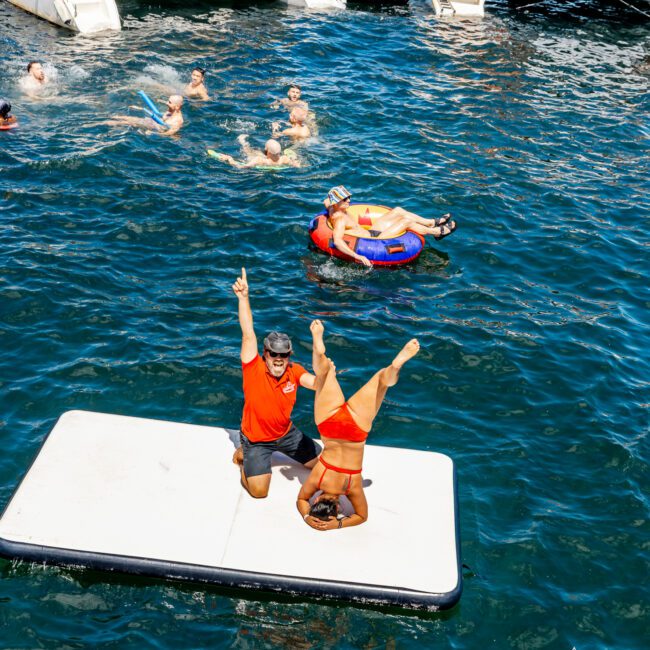 People are enjoying a day out on the water. Two individuals on a floating mat perform yoga poses; one standing with an arm raised and the other in a headstand. Others swim or relax on nearby boats. Banners and logos for "The Yacht Social Club" and Luxury Yacht Rentals Sydney are visible.