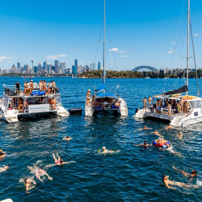 A lively scene on a sunny day with people swimming and enjoying themselves near three docked boats on the water. The Sydney skyline, including the Sydney Harbour Bridge, is visible in the background. Signs on the boats read "The Yacht Social Club Event Boat Charters", and the event appears festive.