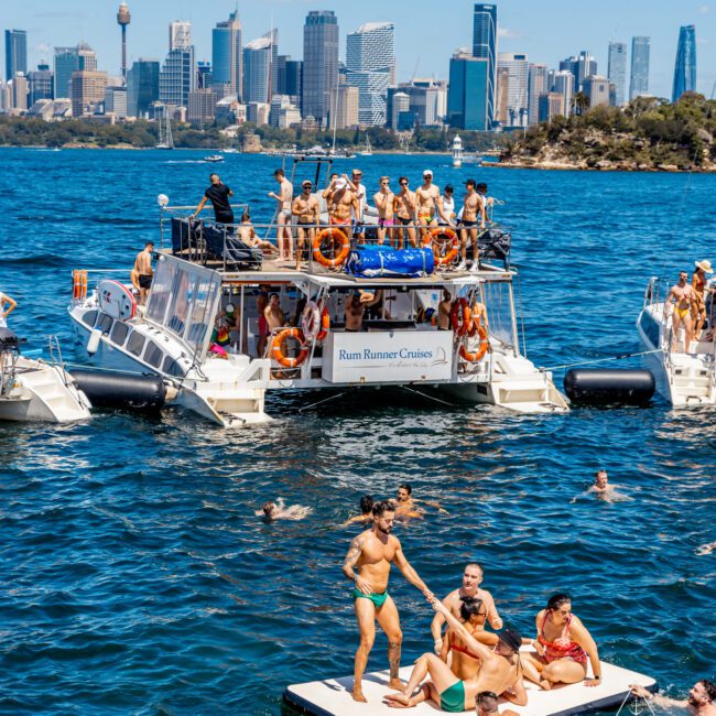 A lively group of people are enjoying a sunny day on and around a boat named "Rum Runner Cruises" in a picturesque harbor. Some individuals are on the boat, while others are on a floating platform in the water. The city skyline is visible in the background, capturing the essence of The Yacht Social Club Sydney Boat Hire.