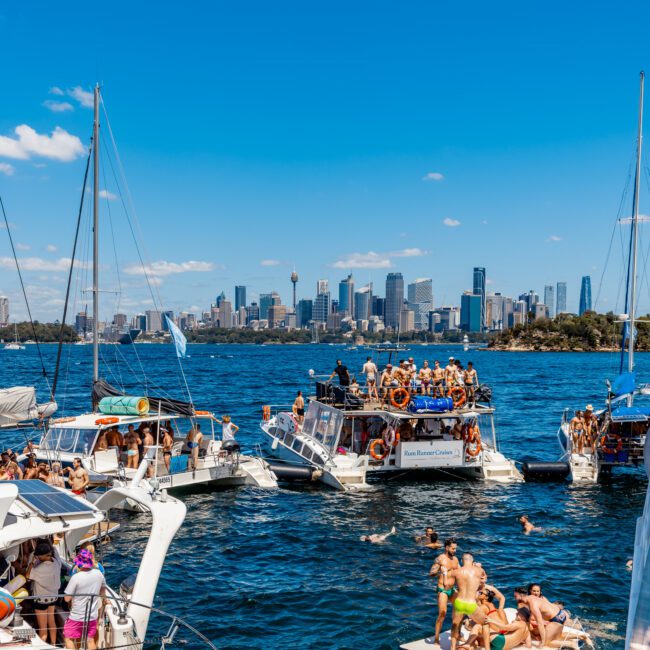 A lively scene on the water, featuring numerous boats with people socializing and enjoying sunny weather. In the background, a city skyline with tall buildings stretches across the horizon under a bright blue sky. The water is a deep blue, and the atmosphere is festive, reminiscent of Boat Parties Sydney The Yacht Social Club.