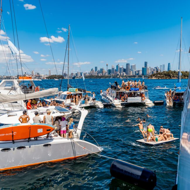 A lively scene with multiple sailboats and motorboats anchored close together on a sunny day. Groups of people are seen socializing on the boats and in the water. The city skyline is visible in the background under a blue sky with scattered clouds, setting the perfect backdrop for The Yacht Social Club Sydney Boat Hire.