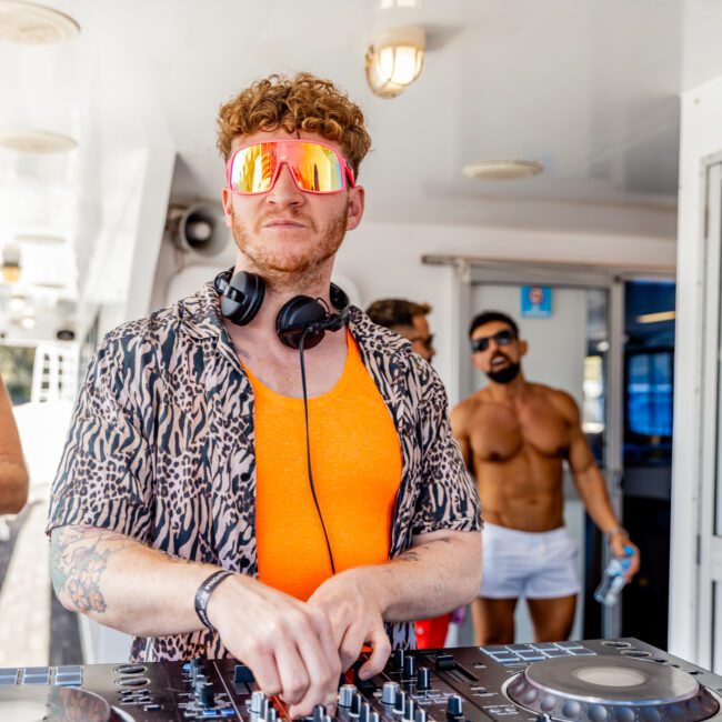 A DJ with curly red hair and large reflective sunglasses, wearing an orange top, is performing on a boat at The Yacht Social Club event. He stands behind a mixer with headphones around his neck. In the background, people in swimwear enjoy the party hosted by Luxury Yacht Rentals Sydney. Logos are visible on the lower corners.