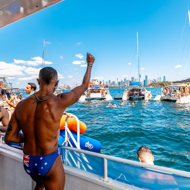 A lively scene on a boat filled with people enjoying a sunny day on the water. A man in the foreground, wearing Australian flag swim briefs, raises his arm in celebration. Other boats from The Yacht Social Club Sydney Boat Hire are in the background, with a cityscape in the distance.