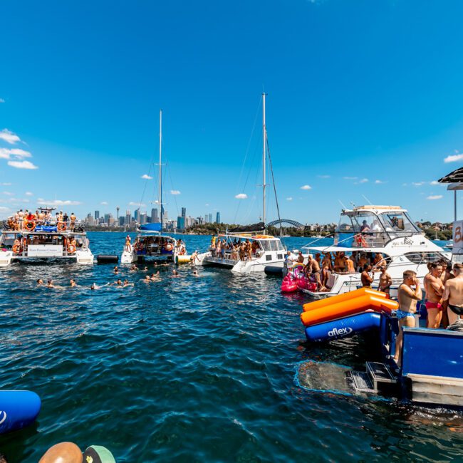 Boats are gathered for a lively event by The Yacht Social Club on a sunny day. People in swimwear are on the boats and in the water, enjoying the festivities. A city skyline is visible in the background under a clear blue sky, while inflatable water toys float around.