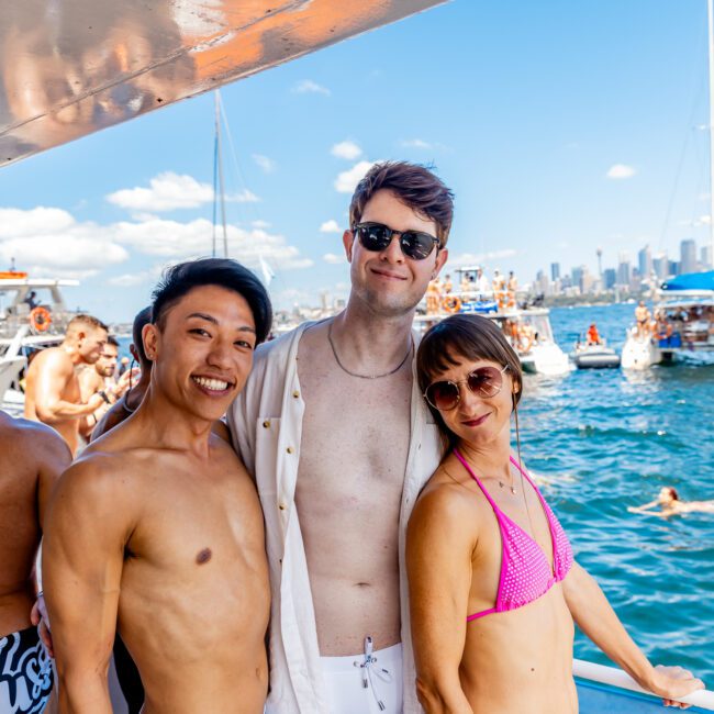 Three people are posing and smiling on a boat during a sunny day, enjoying a delightful outing with The Yacht Social Club. One person is wearing swim trunks, the second is dressed in an unbuttoned shirt and swim trunks, and the third is wearing a pink bikini. Other boats and people are visible in the background on the water.