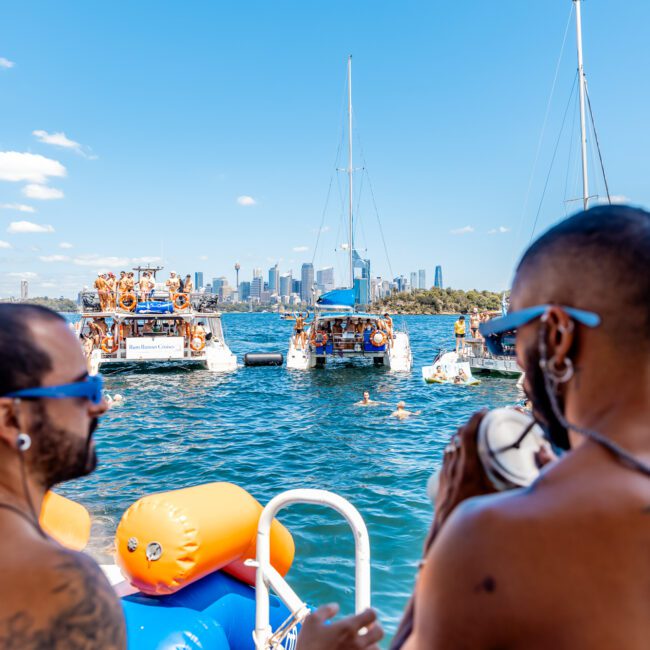 Two people wearing sunglasses stand on a boat, facing away from the camera and looking toward several yachts in the water. Many people are on the yachts, enjoying a sunny day at The Yacht Social Club. In the background, a city skyline is visible under a clear blue sky.