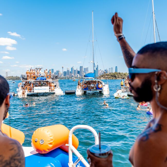 A joyous gathering of people enjoys a sunny day on a yacht in a wide body of water. In the background, two other boats with festive groups are seen, with a city skyline under a clear blue sky. One individual in the foreground raises a hand, holding a drink. Boat Parties Sydney The Yacht Social Club add to the fun.