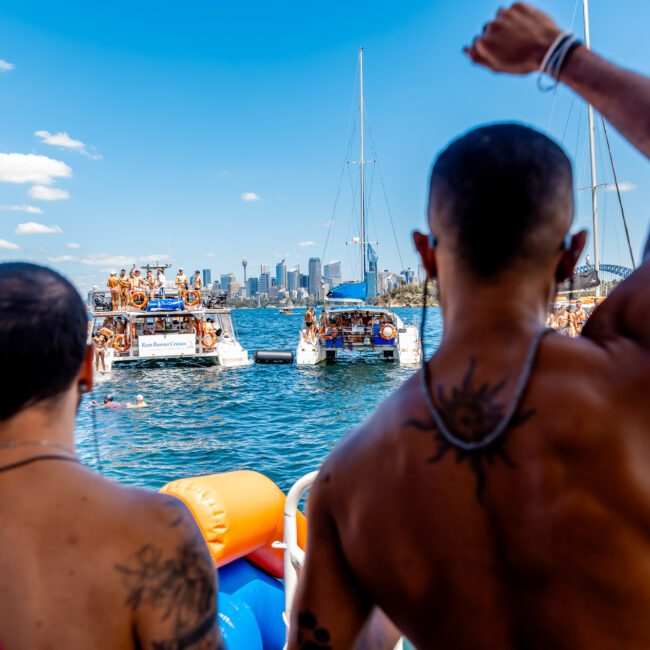 People enjoying a lively boat party on the water near a city skyline. The weather is sunny, and the group appears cheerful and energetic, with one person raising their arm in celebration. Sailboats are anchored nearby with more partygoers aboard. Logos "GAYM" and "The Yacht Social Club Sydney Boat Hire" are visible.