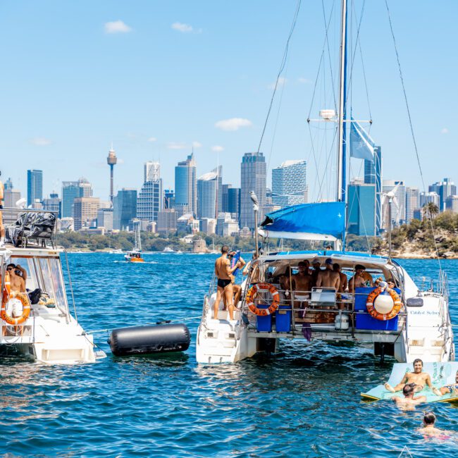 A group of people enjoying a sunny day on two yachts anchored close together in a vibrant blue bay. Some are swimming in the water, others lounging on the boats. The city skyline with tall buildings and a tower is visible in the background, as part of The Yacht Social Club Sydney Boat Hire experience.