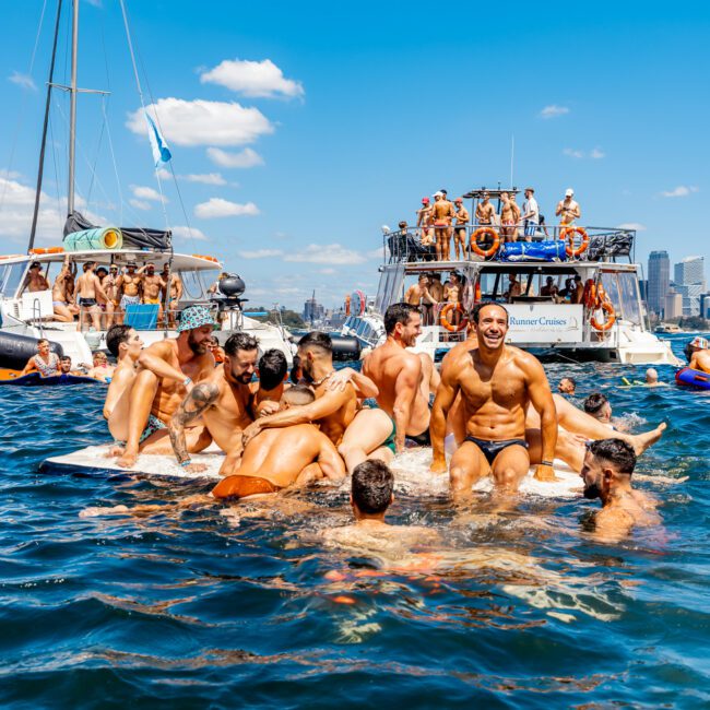 A group of people are gathered on a floating mat in a large body of water, engaging in a cheerful activity. Nearby boats from The Yacht Social Club are anchored with more people onboard. The sky is clear and sunny, and the distant city skyline is visible in the background.