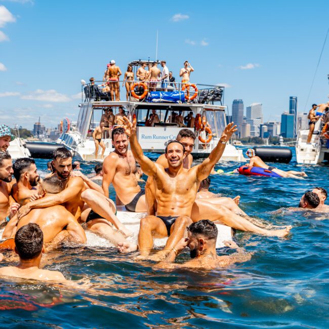 A group of people enjoying a sunny day on the water, surrounding boats in the background. Some are on an inflatable float, waving and smiling, while others swim nearby. Skyscrapers are visible in the distance against a clear blue sky. Logos "The Yacht Social Club Sydney Boat Hire" and "Yacht Social Club" are in the bottom corners.