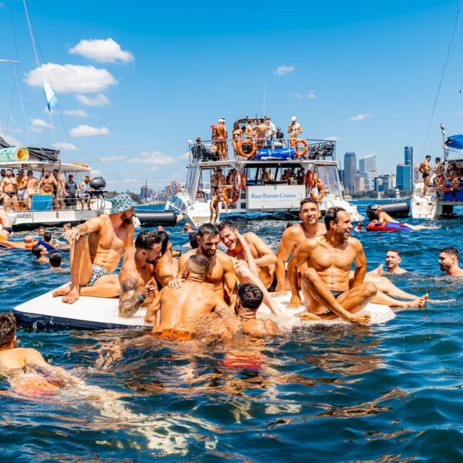 A lively group of people enjoys a sunny day on the water, swimming and lounging on a floating platform. Several boats from The Yacht Social Club are anchored nearby, with a city skyline visible in the background under a clear blue sky.