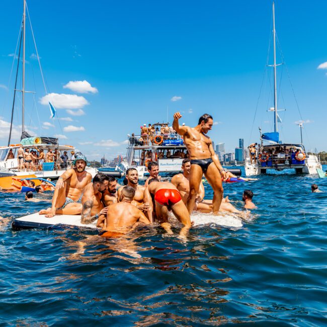 People enjoying a sunny day on a lake with multiple yachts and boats in the background. A group of men in swimwear are having fun on a floating platform in the water, reminiscent of The Yacht Social Club's lively boat parties in Sydney. The scene boasts clear blue skies and city buildings visible in the distance.