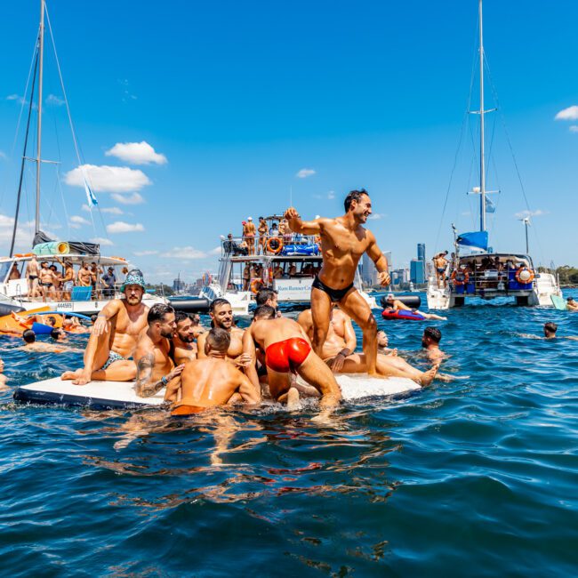 A group of people on a floating platform enjoying themselves in a large body of water surrounded by boats. Many are in swimwear. The sky is clear with a few clouds, and city buildings are visible in the background. The atmosphere is lively and festive, reminiscent of The Yacht Social Club's events.