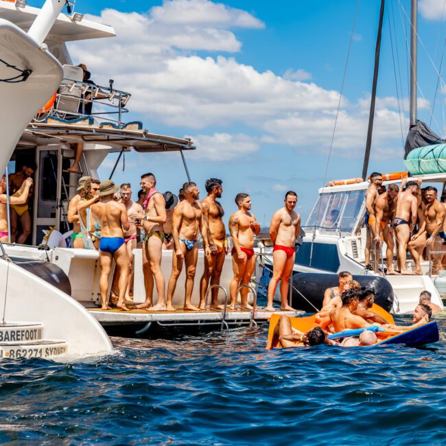 People are enjoying a sunny day on boats and in the water, with some sunbathing on decks and others swimming or floating on inflatable mats. Several sailboats are anchored close together. It's a perfect setting for The Yacht Social Club Sydney Boat Hire, under a bright and clear sky with a few clouds.