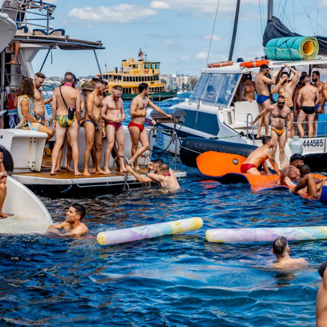 A lively boat party hosted by The Yacht Social Club is taking place with numerous people in swimsuits on the deck of a yacht and in the water. Several flotation devices, including a large inflatable orange unicorn, add to the fun. Other boats are moored nearby under a partly cloudy sky.