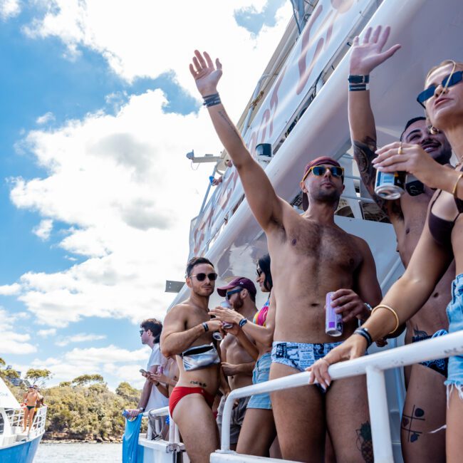 A group of people in swimwear are on a boat, dancing and enjoying a sunny day. One man raises his arm, holding a drink. The Yacht Social Club Event Boat Charters are docked near a scenic shoreline with trees visible in the background. Branded logos appear on the lower portions of the image.