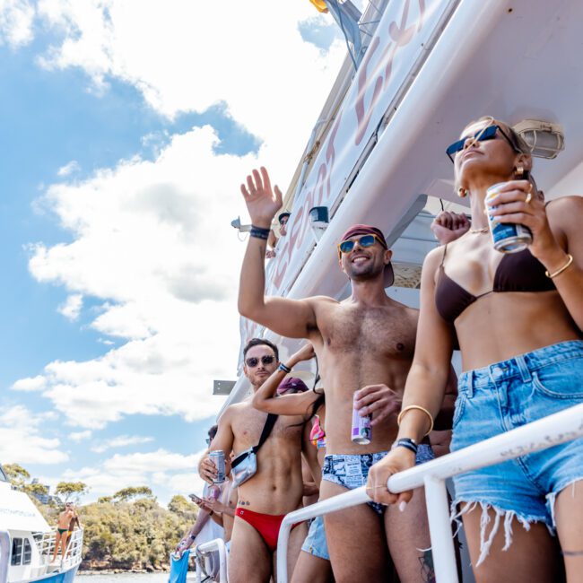 A group of people on the deck of a yacht are enjoying a sunny day. Dressed in swimwear and sunglasses, they hold drinks while waving and smiling at the camera. The Yacht Social Club's boat is set against a blue sky with some clouds, and another boat is visible in the background.