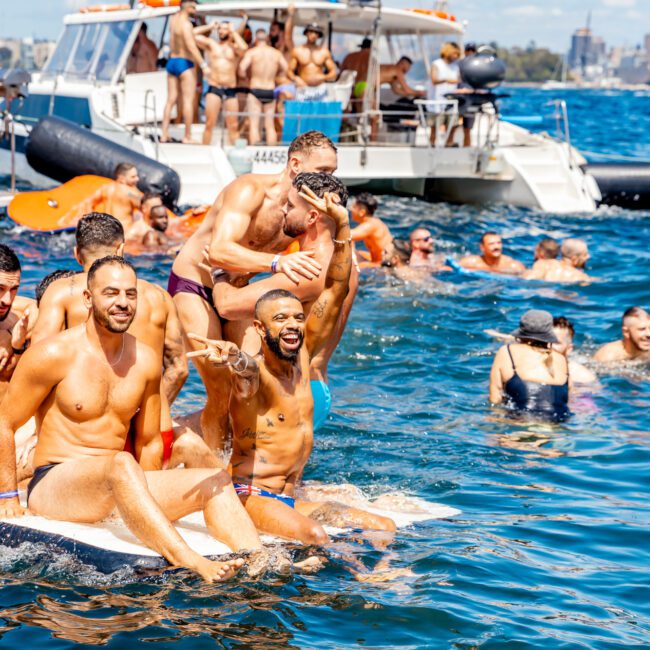 A vibrant group of shirtless men enjoys a day on the water, some on a raft and others swimming around. In the background are boats and more people. The sky is clear and the water is a deep blue, creating a lively and festive atmosphere. Logos of Sydney Harbour Boat Hire The Yacht Social Club are visible.