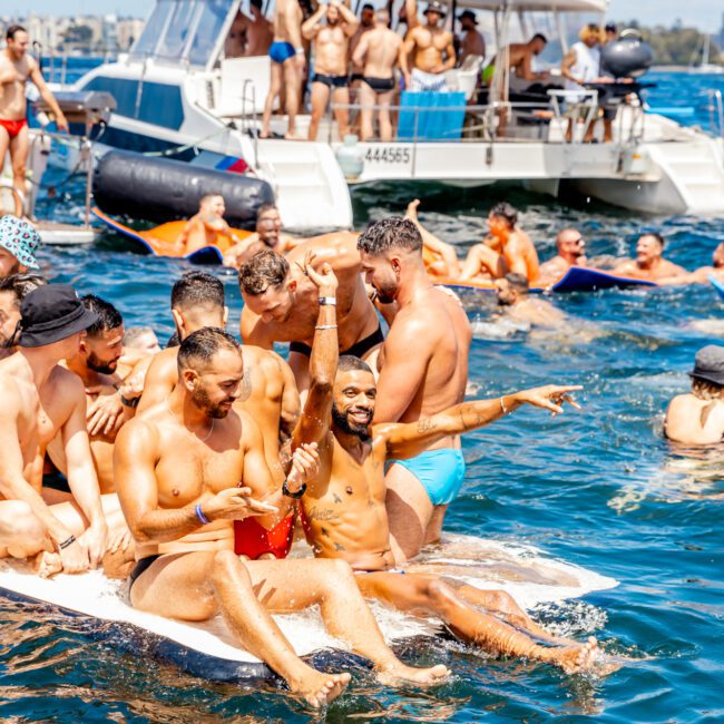 A group of men in swimsuits enjoy a sunny day on the water. Some sit on the edge of a floating platform, smiling and making gestures, while others swim around or relax on a nearby boat. The background features more revelers from The Yacht Social Club Sydney Boat Hire, enjoying an unforgettable outing.