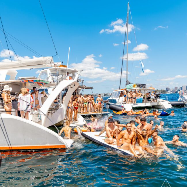 A lively group enjoys a sunny day on boats and in the water. Dozens gather on yachts and inflatable platforms, courtesy of The Yacht Social Club Event Boat Charters, smiling and conversing. The sky is mostly clear, with distant city buildings visible under the bright blue sky.