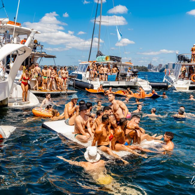 A lively summer scene on the water with multiple boats and yachts filled with people enjoying the sun. Groups of individuals are swimming, lounging on a floating platform, and using inflatable rafts, creating a festive atmosphere under a clear blue sky during The Yacht Social Club Event Boat Charters.