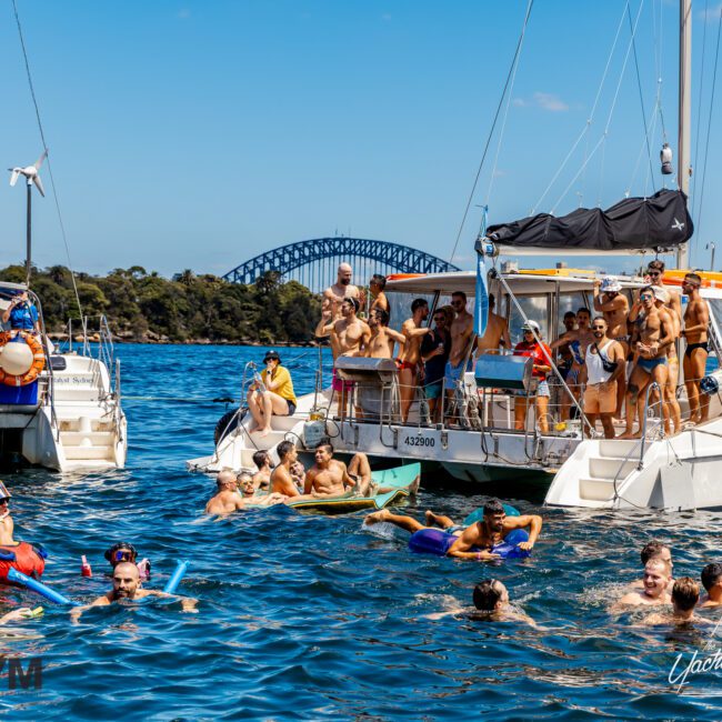 A large group of people enjoys a sunny day on the water, swimming and lounging on inflatable devices near two boats. The Sydney Harbour Bridge is visible in the background under a clear blue sky. A festive atmosphere pervades the scene, courtesy of The Yacht Social Club Sydney Boat Hire.