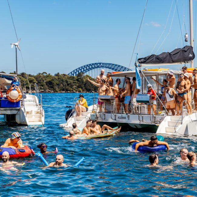 A group of people enjoying a sunny day on and around two anchored boats in a vibrant blue body of water. Some are swimming with flotation devices, while others socialize on the boats. A large bridge and lush greenery are visible in the background, setting the scene for The Yacht Social Club event boat charters.