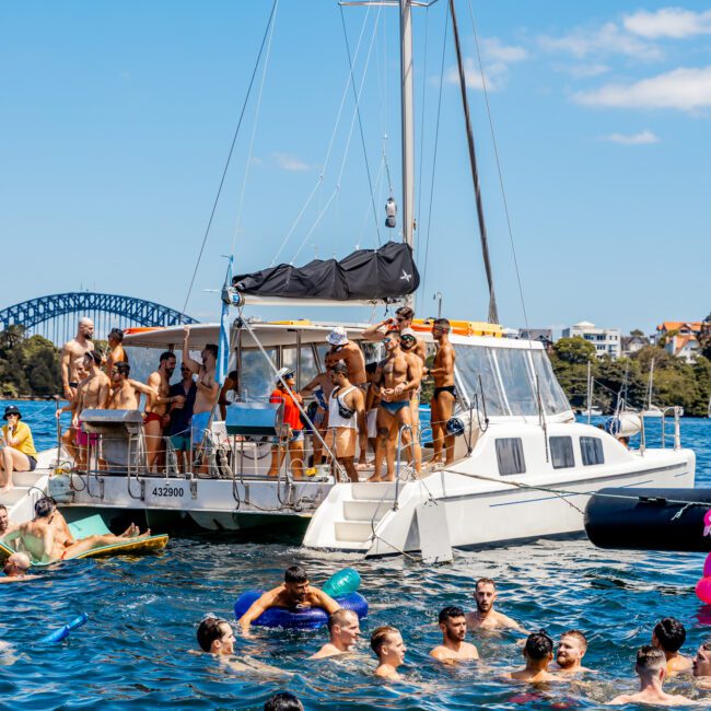 A group of people having fun on a catamaran and swimming in the water around it on a sunny day. The scene includes a mix of people on the boat and in the water, with a bridge and cityscape visible in the background. Logos for "AYM" and "Yacht Social Club" are on the bottom corners, promoting The Yacht Social Club Event Boat Charters.