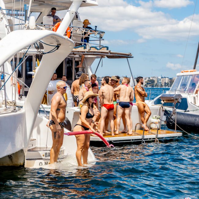 A lively group of people in swimwear are enjoying time on and around the deck of a white yacht. Some are standing on a smaller platform attached to the yacht, while others are on the main deck. The sea and a clear blue sky are in the background, showcasing The Yacht Social Club Event Boat Charters.